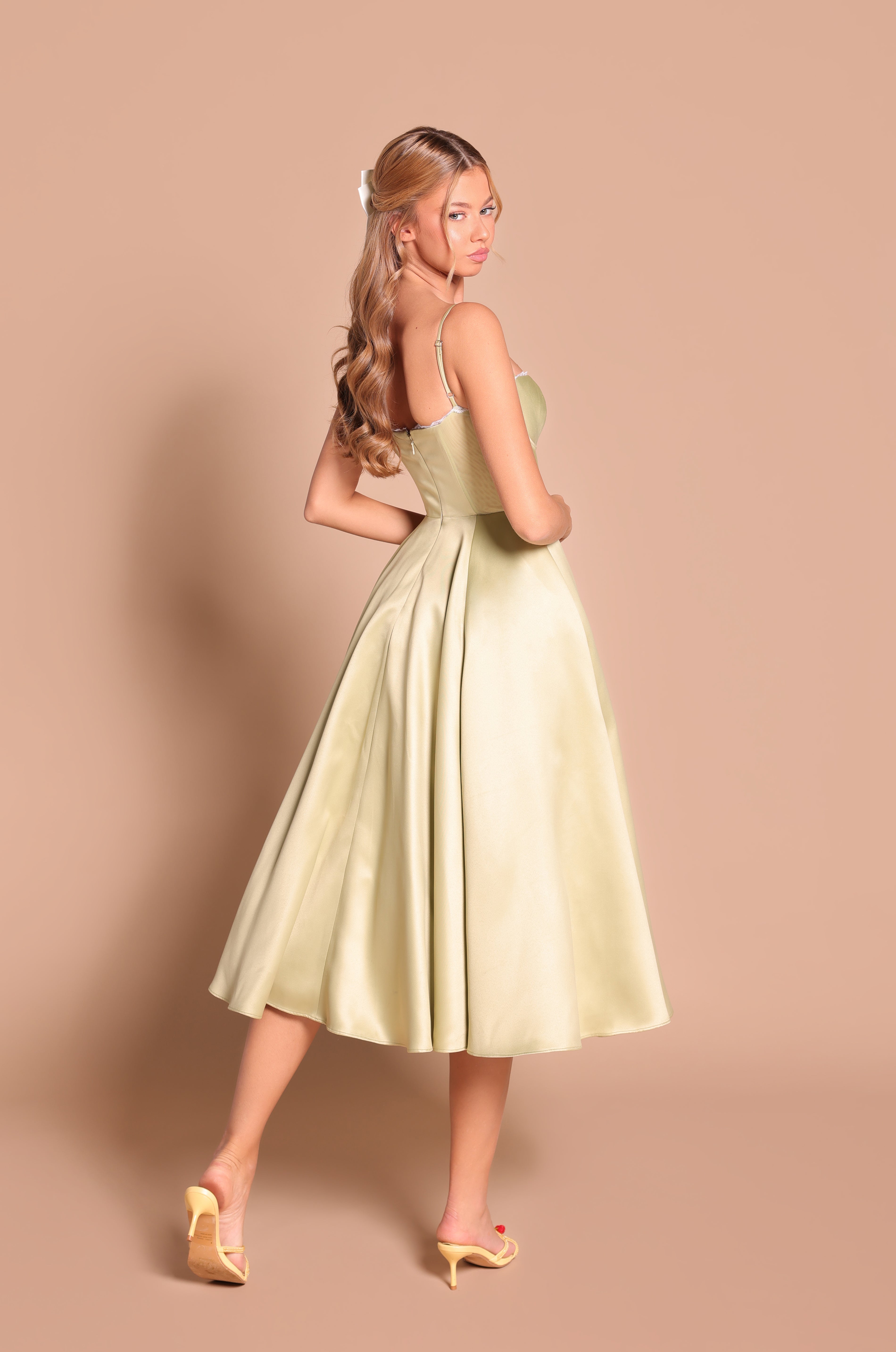Woman with long wavy hair wears the Olive Green Satin Midi Wedding Guest Dress: Olivetta and yellow heels, standing against a beige background, turned slightly to the side.